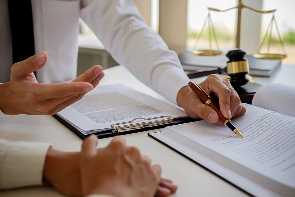 In a law firm office, a legal counsel presents a signed contract to the client, emphasizing justice and law. The scene features a gavel on the table symbolizing legal proceedings, with a law team having a meeting in the background, discussing legal matters.