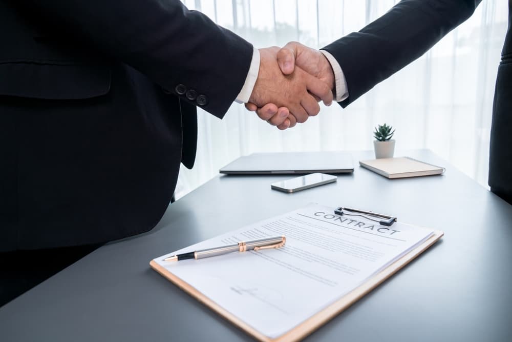 Two professionals successfully close a business deal with a firm handshake, sealing their partnership agreement. In the close-up shot, their hands clasp firmly, with a legal document visible in the background, symbolizing the formalization and mutual commitment to the agreement.

