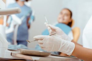 Dentist wearing gloves preparing instruments while a patient waits in the chair during a dental exam.