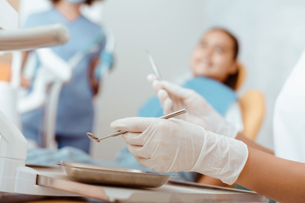 Dentist wearing gloves preparing instruments while a patient waits in the chair during a dental exam.